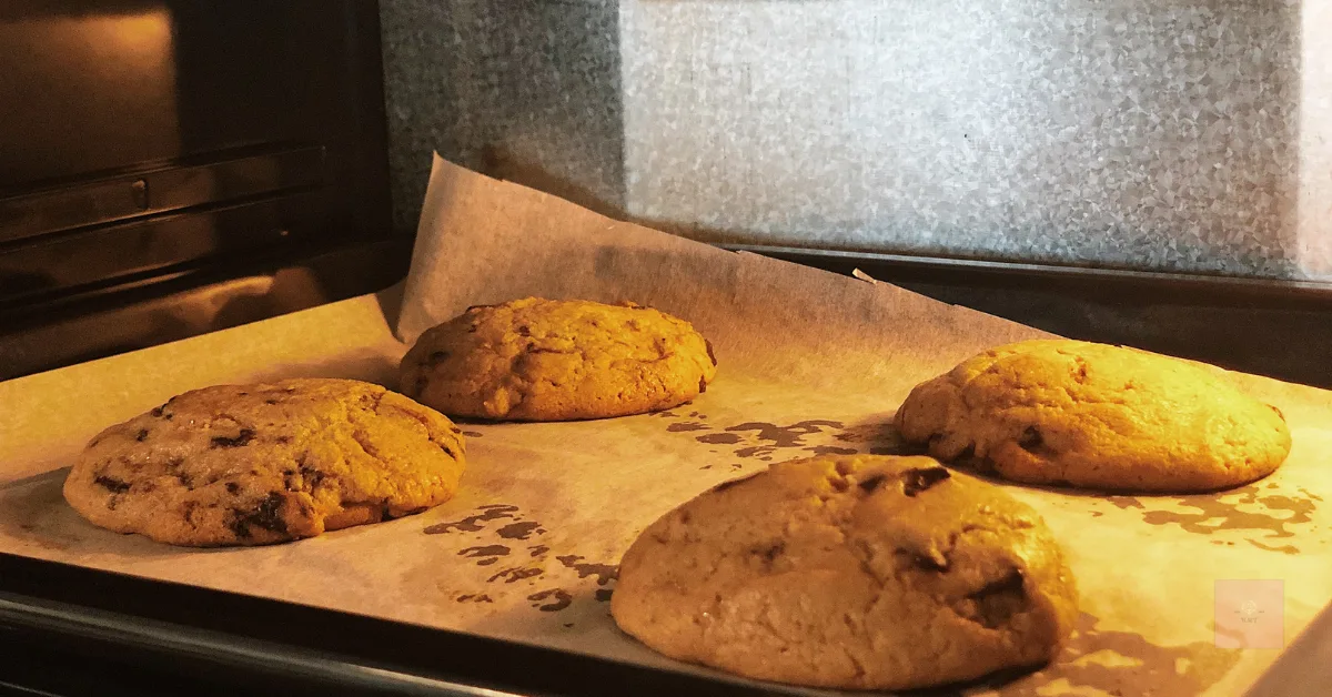 Four golden brown cookies baking on a parchment-lined tray inside an oven, lit warmly from within.